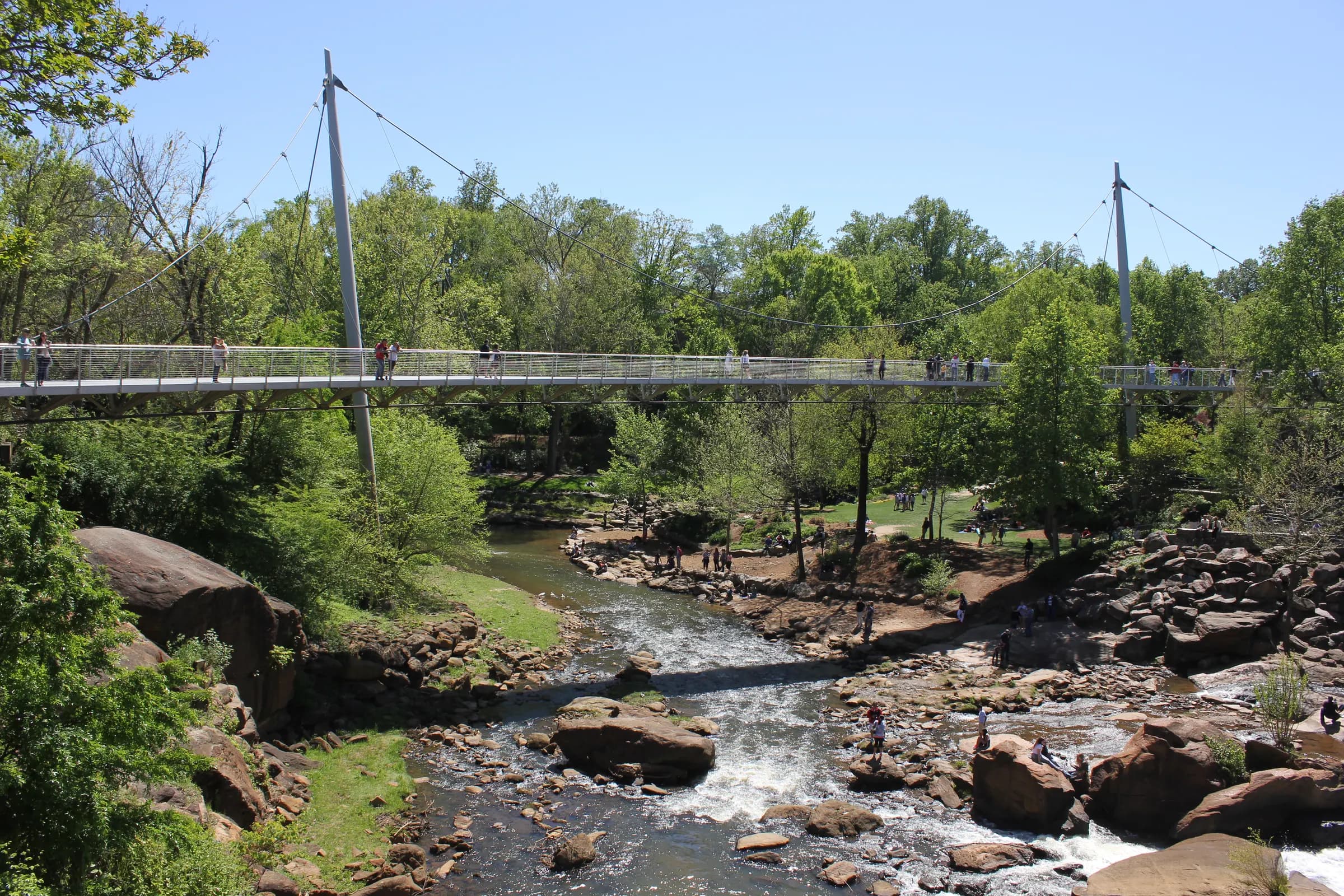The Liberty Bridge curving over the Reedy River falls in Falls Park, Greenville, under a clear spring sky.