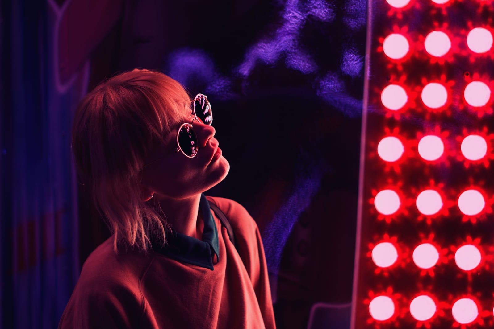 A person in round sunglasses looking up, lit in red and violet light beside a panel of red light-therapy lamps.
